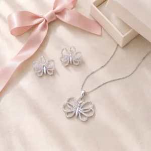 The Soft Wing Set laid out on linen: butterfly earrings and the butterfly pendant on its box chain, with a pink satin bow and gift box